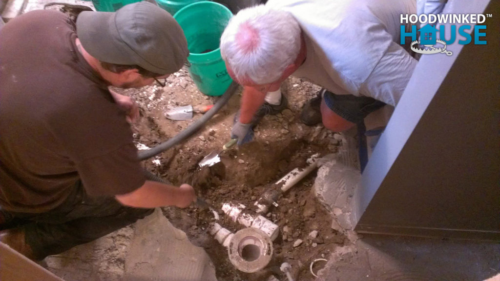 Two men dig underground pipes out of a basement floor.