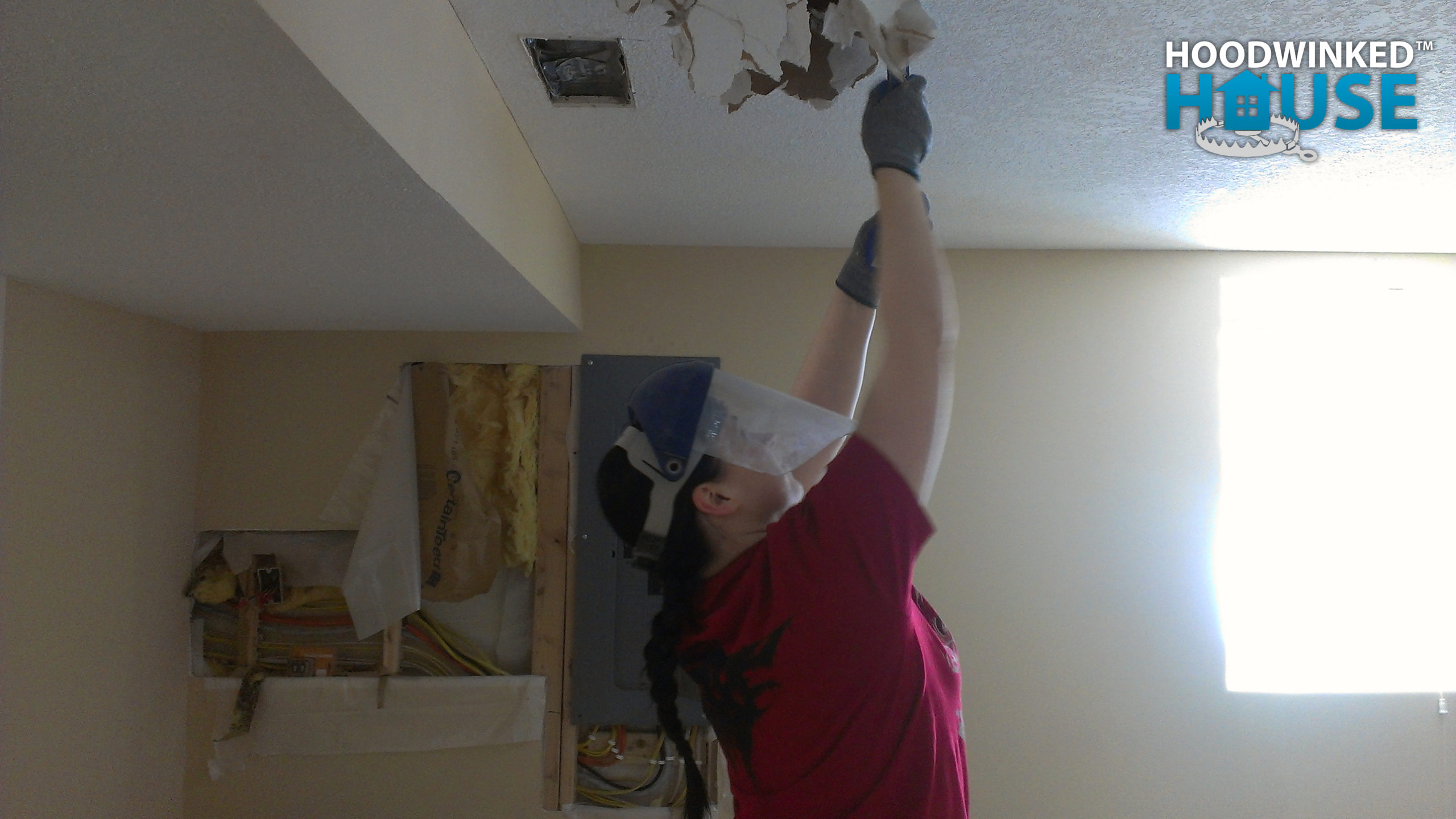 A woman wearing a face shield and gloves uses a prybar to demolish drywall from a bedroom ceiling.