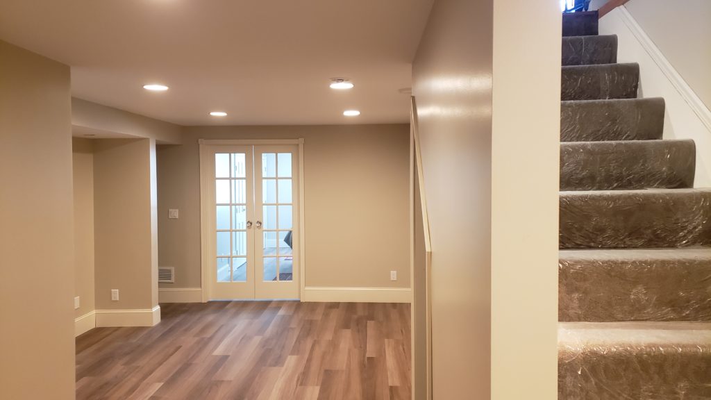 A finished basement without furniture, showing recessed lighting, wood grain vinyl flooring, and double French glass doors.