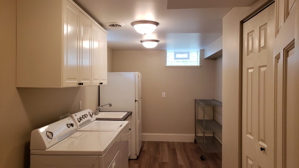 A finished basement laundry room with wood plank vinyl flooring, white laundry machines, white cabinetry, laundry tub, a refrigerator, and a commercial rolling shelf.