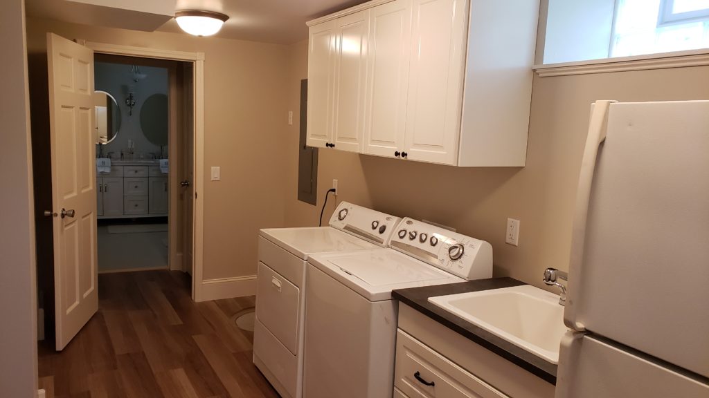 A finished basement laundry room with wood plank vinyl flooring, white laundry machines, white cabinetry, laundry tub, and a refrigerator.