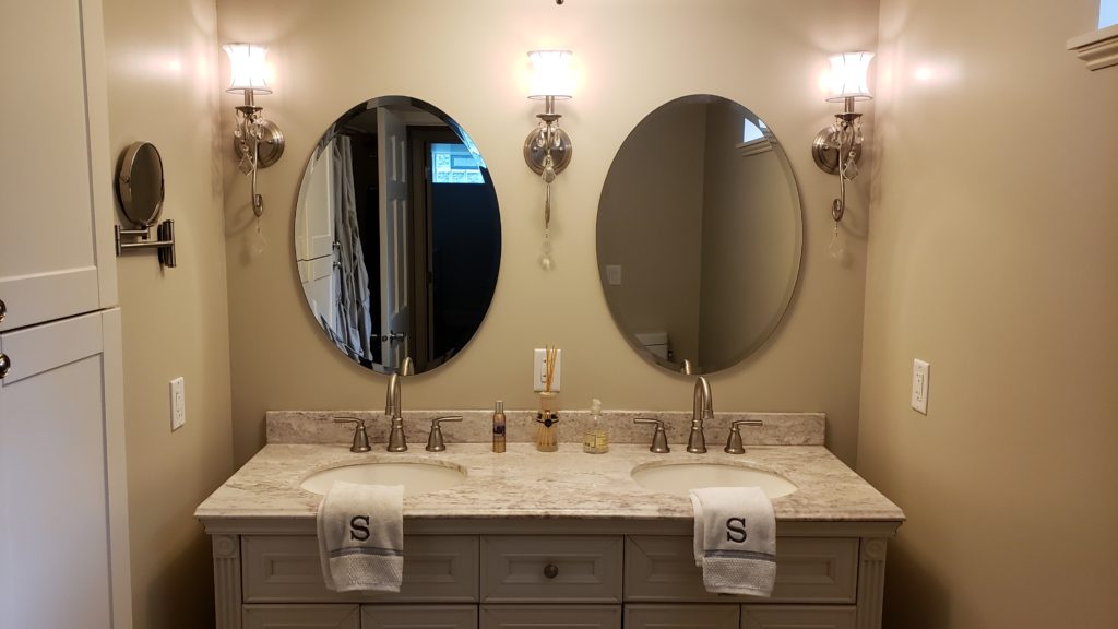 A bathroom vanity with a marble top, two sinks, two oval mirrors, ornate sconce lighting, and two monogrammed towels.