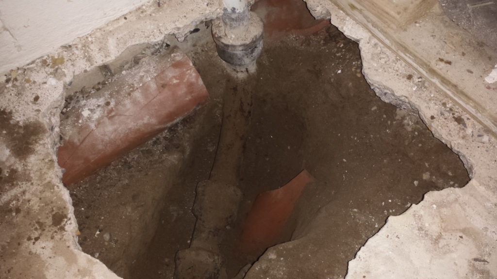Excavating a basement floor to expose a pipe junction reveals that legacy clay drain tile had been broken to make way for the pipe.
