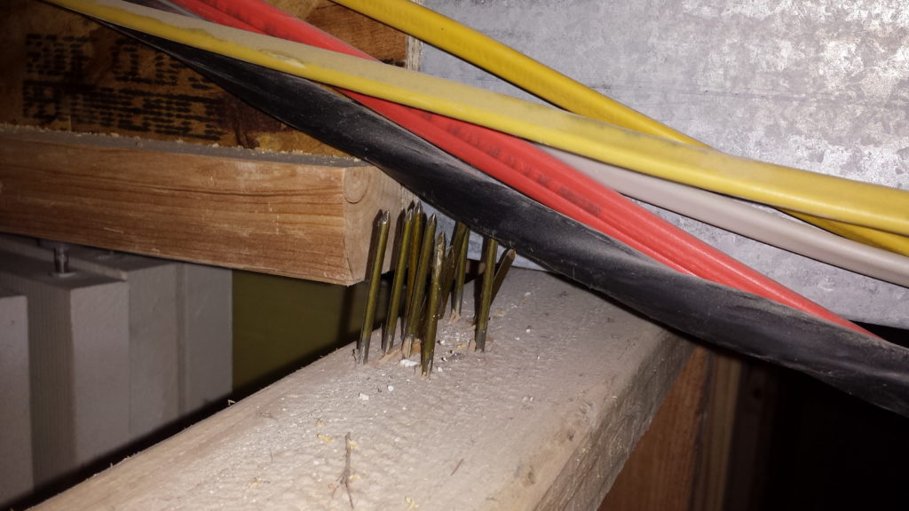 Stud wall framing above a ceiling shows many sharp framing nails touching some large electrical wires.