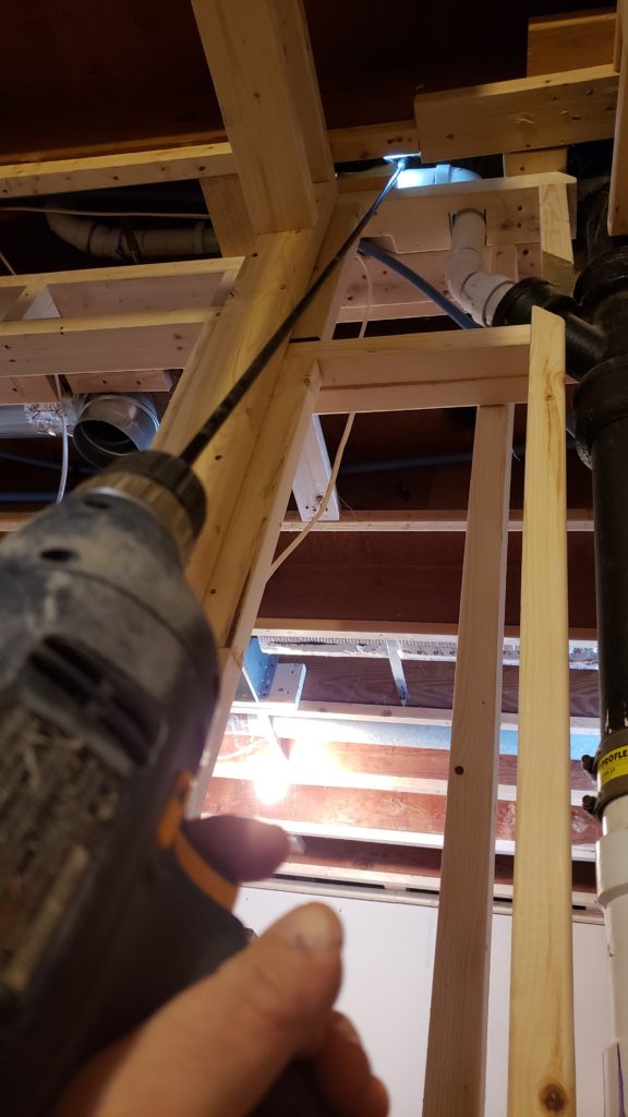 Using a long drill bit to drill a hole between two pipes in a basement ceiling.