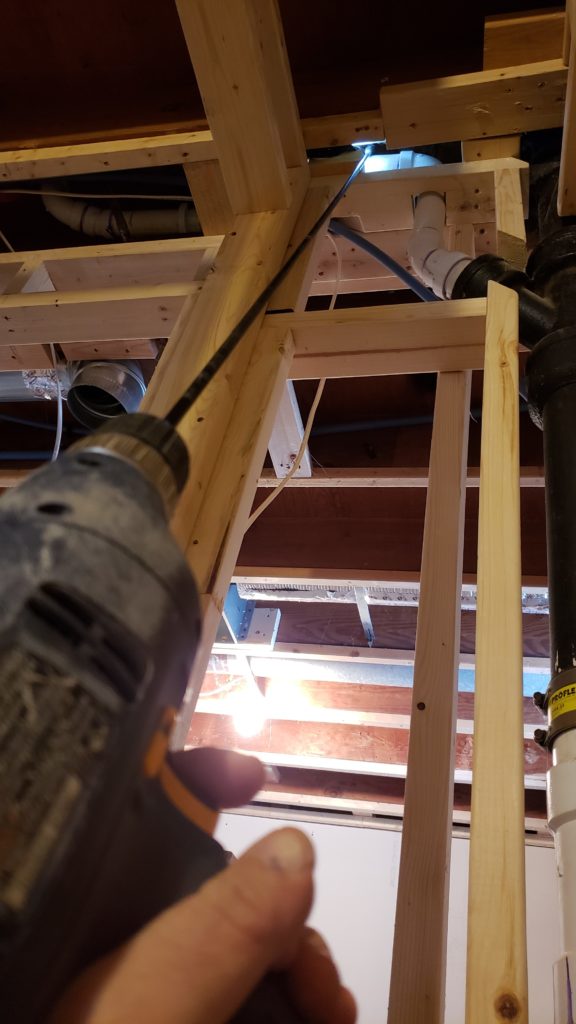 Using a long drill bit to drill a hole between two pipes in a basement ceiling.