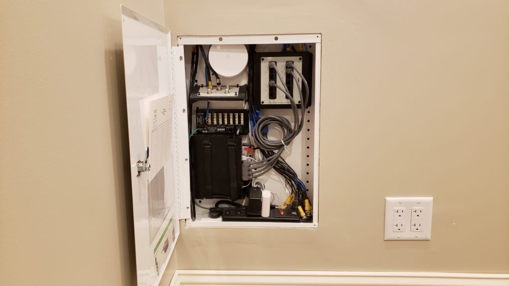 A structured media enclosure inset into a wall with the door open showing patch panels for telephone, cable TV, a household computer network, security camera system, as well as a modem and Wi-Fi router.