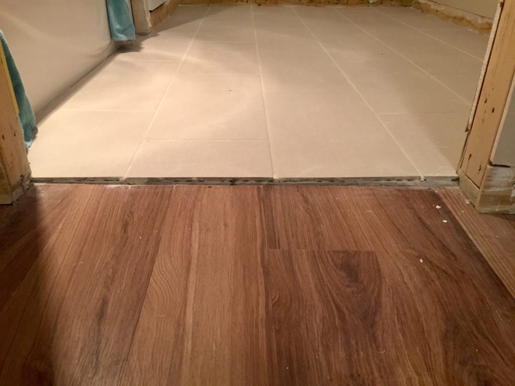 A wood grain vinyl floor meets a bathroom tile floor, but it missing a transition between the two.