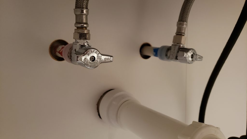 Under a laundry tub, hot and cold pipes are shown with shutoff valves. The water pipes and drain pipe protrude from perfect holes in the back of the cabinet.